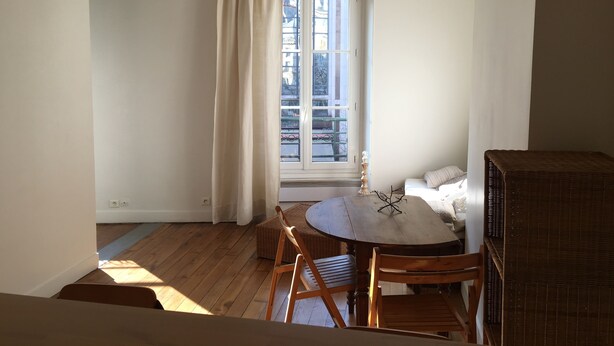 Een kamer met een houten vloer, een ronde tafel met stoelen, een raam met gordijnen, en een rieten bank.