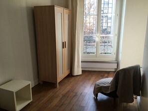 A room with a wooden wardrobe, a window with a view of buildings, and a chair. - Charming, bright 2-room apartment near the Luxembourg Gardens (Paris)