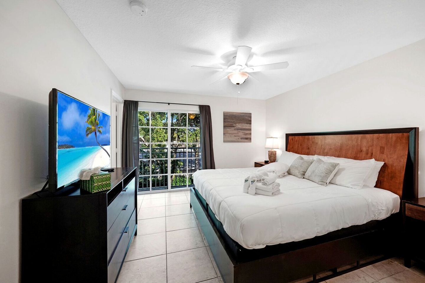 A hotel room with a bed, a television, a ceiling fan, a nightstand, and a view of the outdoors.
