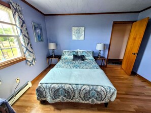 A bedroom with a bed, two bedside tables, a window with curtains, and a door. - Scenic Coastal Cottage with Ocean Views Near Acadia National Park in Lamoine, Maine (Lamoine Point)