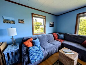 A cozy living room with a grey sofa, a small side table with a lamp, and a window with a view of greenery. - Scenic Coastal Cottage with Ocean Views Near Acadia National Park in Lamoine, Maine (Lamoine Point)