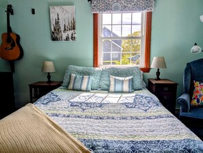 Une chambre à coucher avec un lit, deux tables de chevet, une guitare, une fenêtre donnant sur un beau paysage et une chaise.