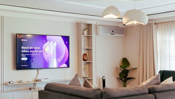 Un saló modern amb televisió plana que mostra un anunci de YouTube, un sofà gris, una planta i un aparell d'aire acondicionat blanc.