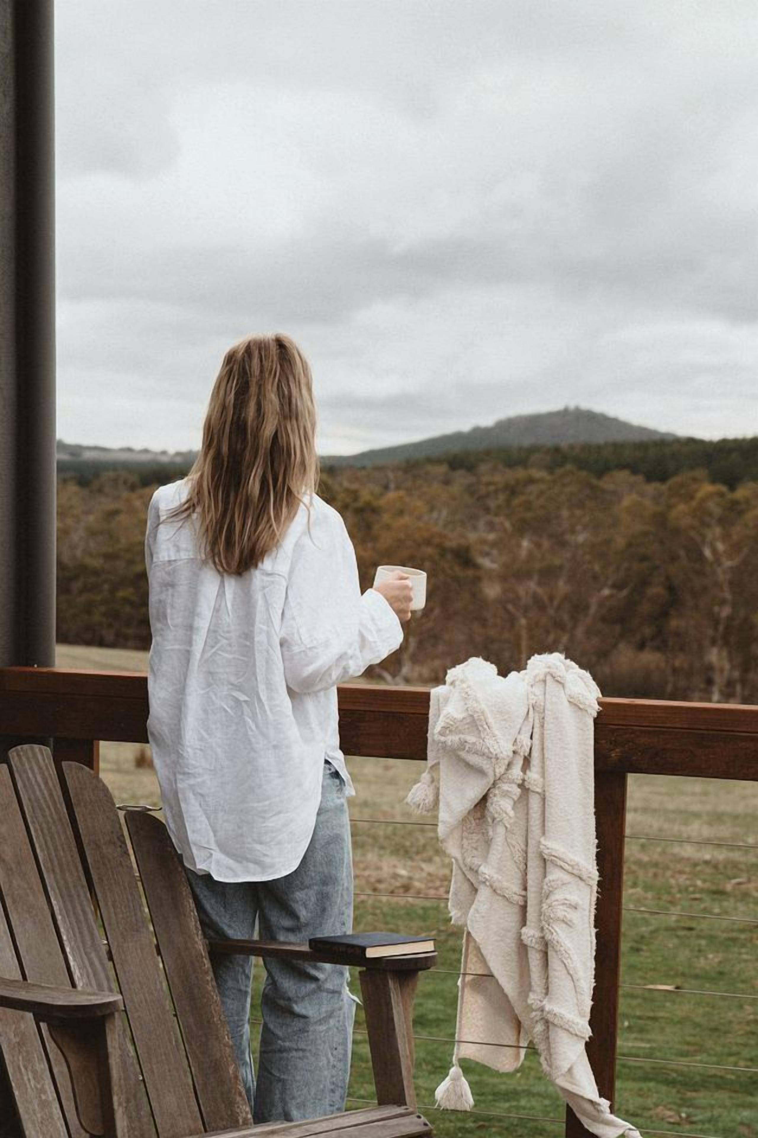 Una persona sentada en una silla de madera, sosteniendo una taza, con vistas a una ladera boscosa.