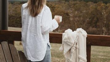 Una persona sentada en una silla de madera, sosteniendo una taza, con vistas a una ladera boscosa.