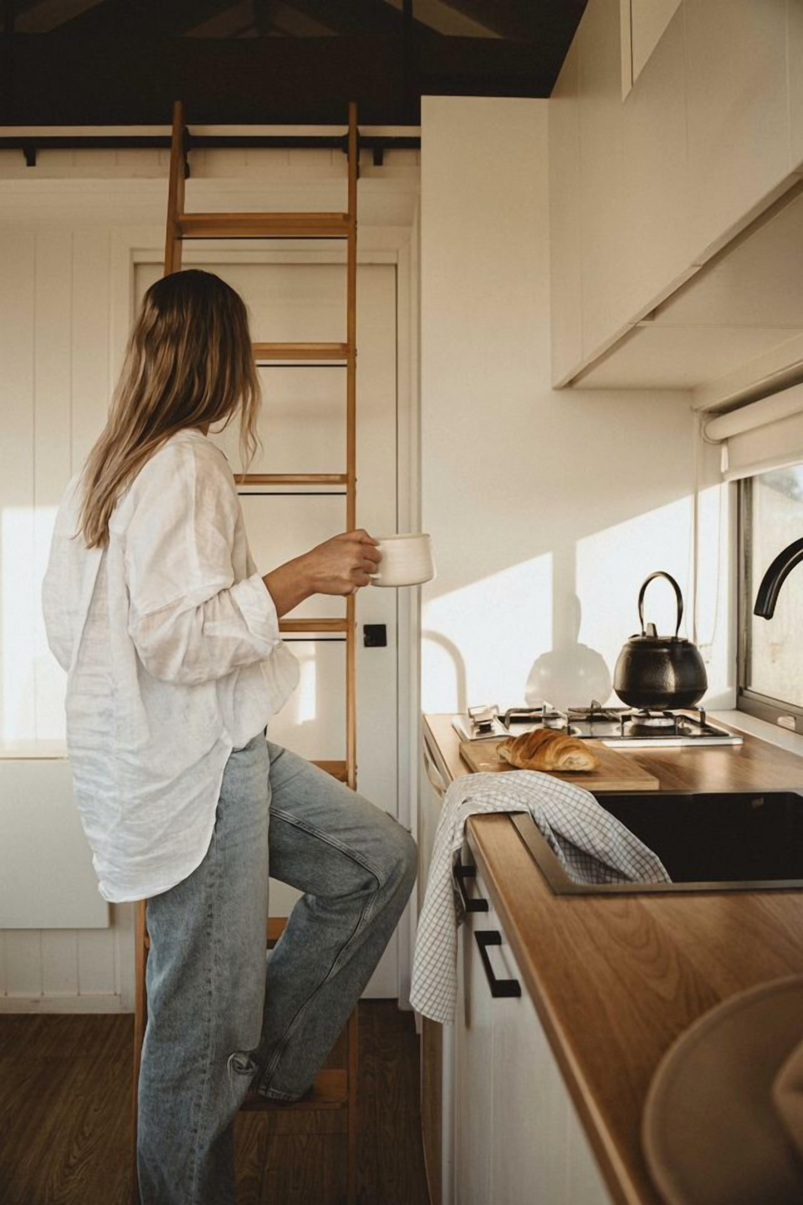 Una persona de pie en una cocina con una escalera de madera, una tetera y un croissant sobre la encimera.