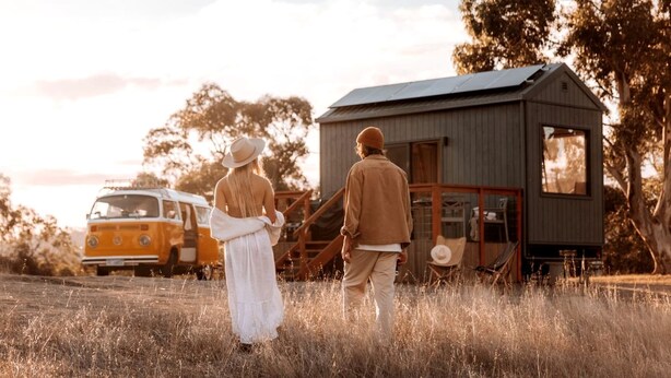 Una pareja parada en un campo, con un coche antiguo y una cabaña pequeña al fondo.