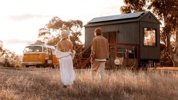 A couple standing in a field with a vintage car and a small cabin in the background.