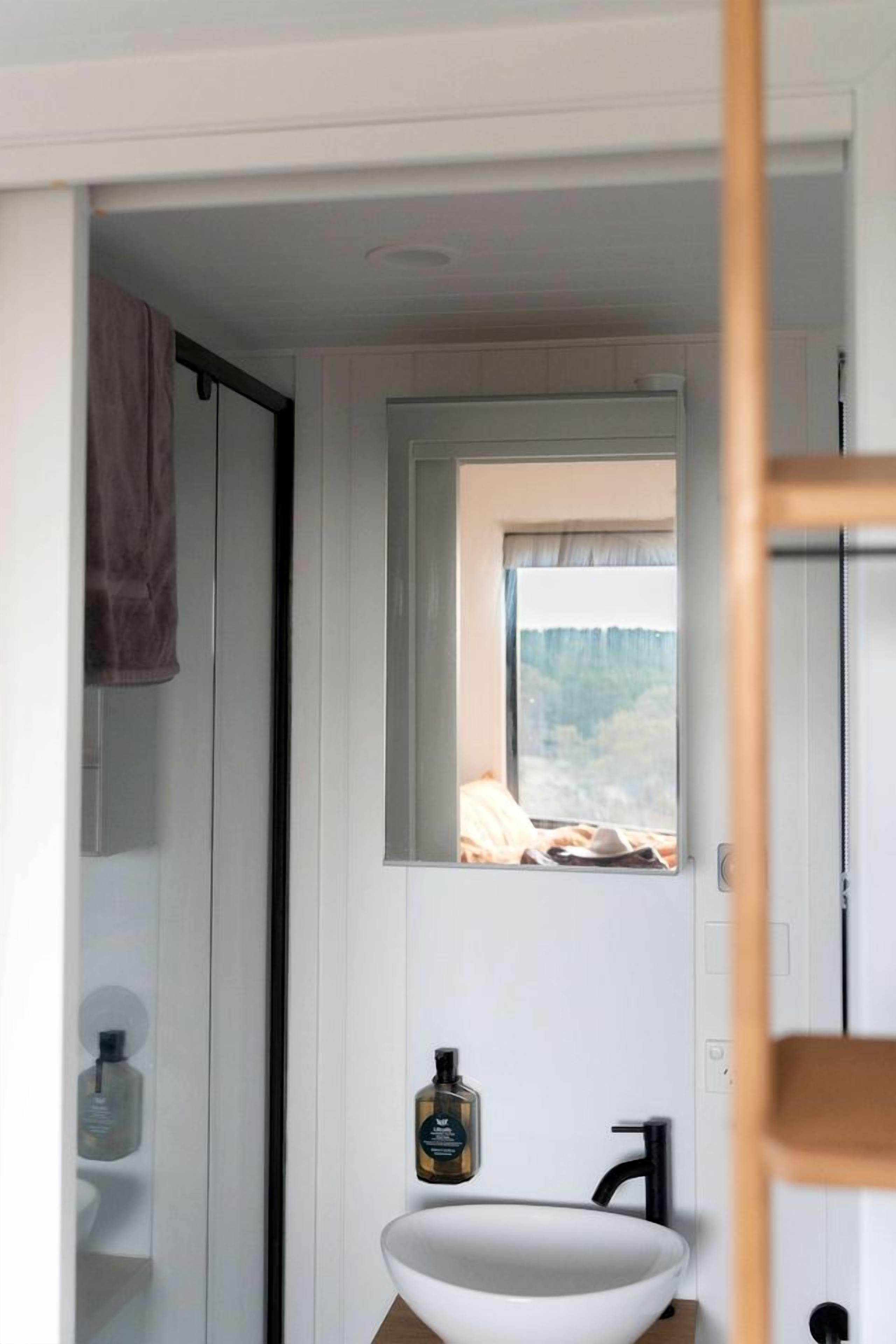 A bathroom with a white sink, a mirror, and a window with a view of a bed and a blanket.
