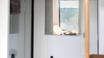 A bathroom with a white sink, a mirror, and a window with a view of a bed and a blanket.