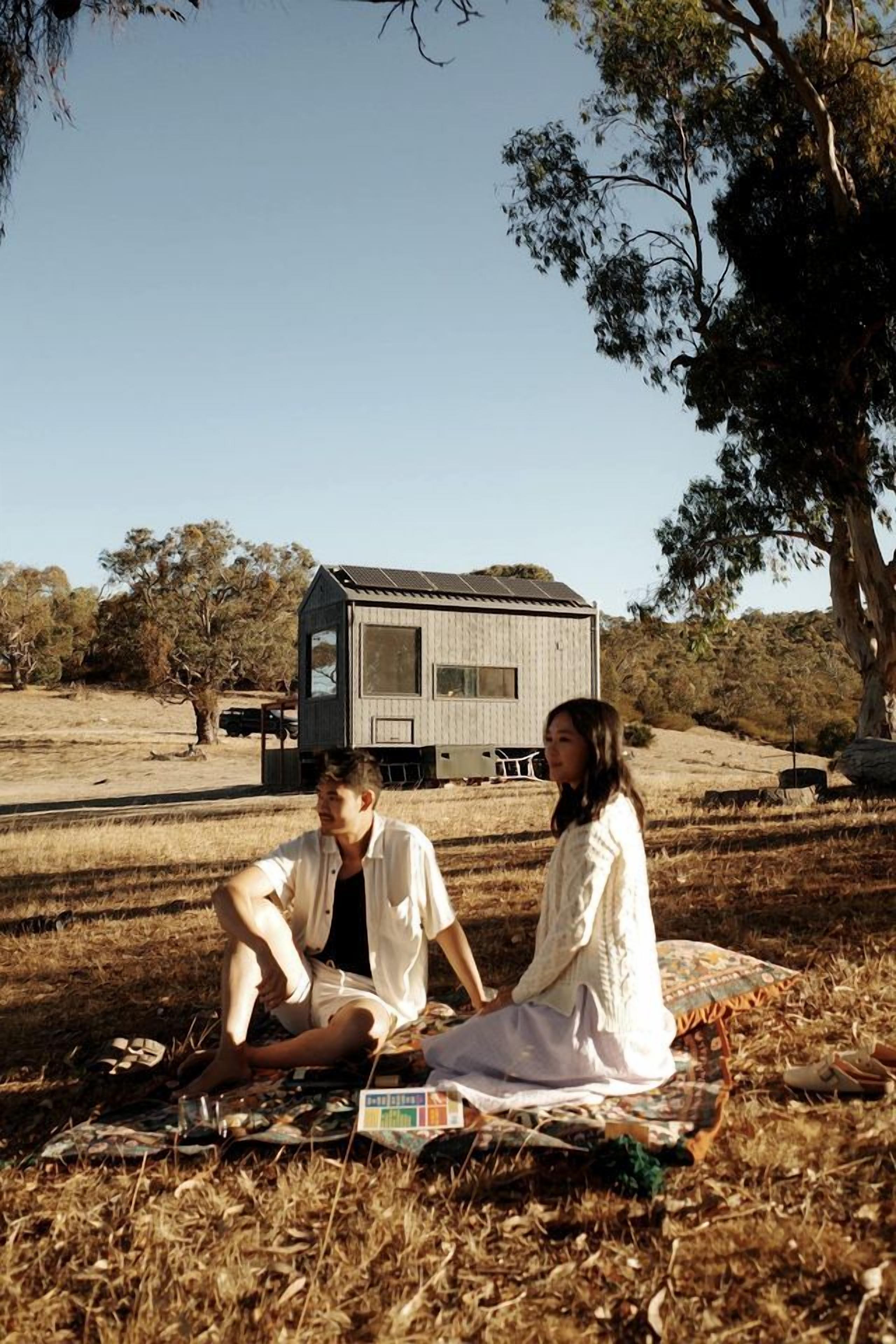 Two people sitting on a blanket outdoors with a small wooden structure in the background.