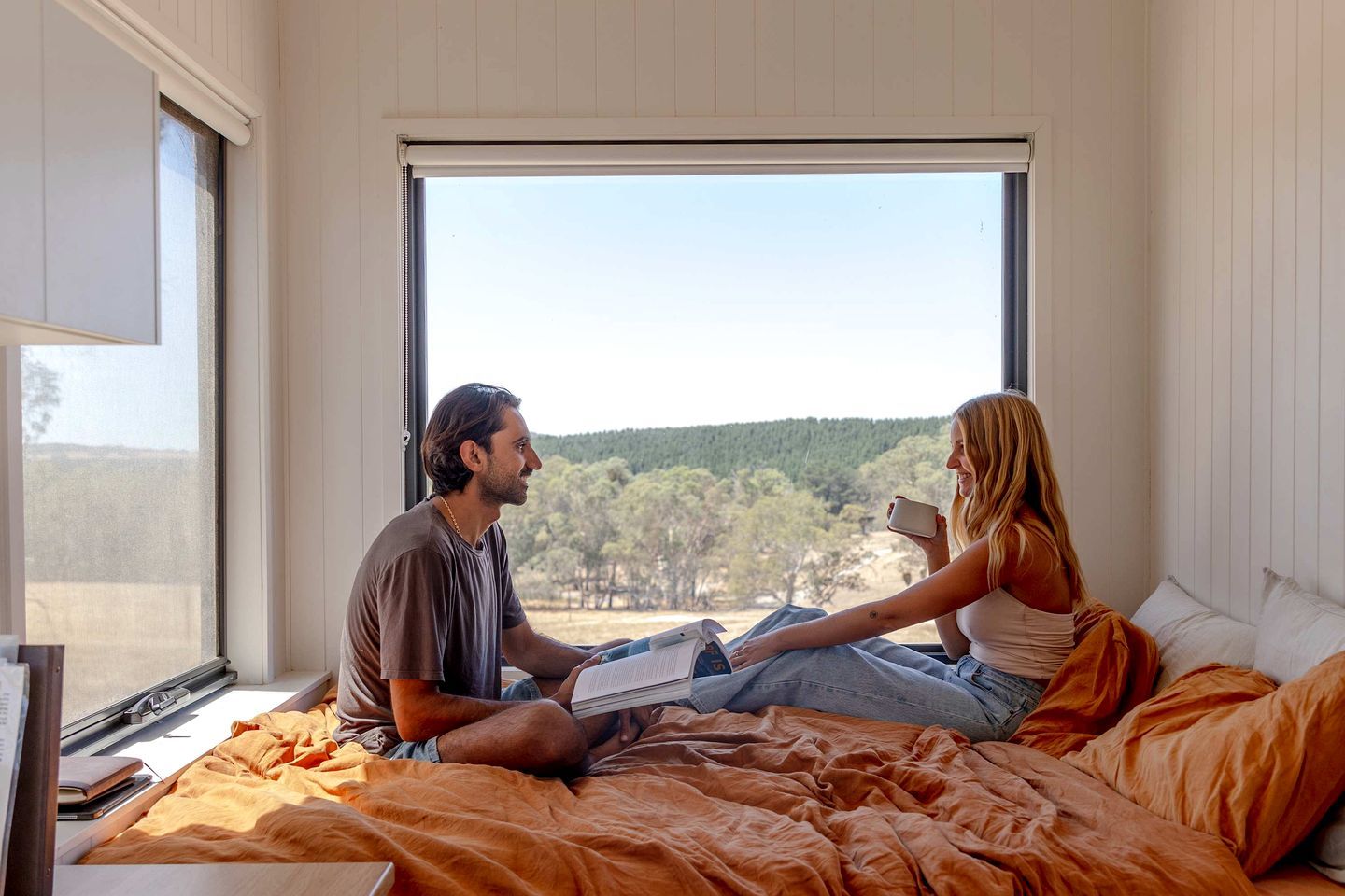 A bedroom with a large window, a bed with orange bedding, and a person reading a book.