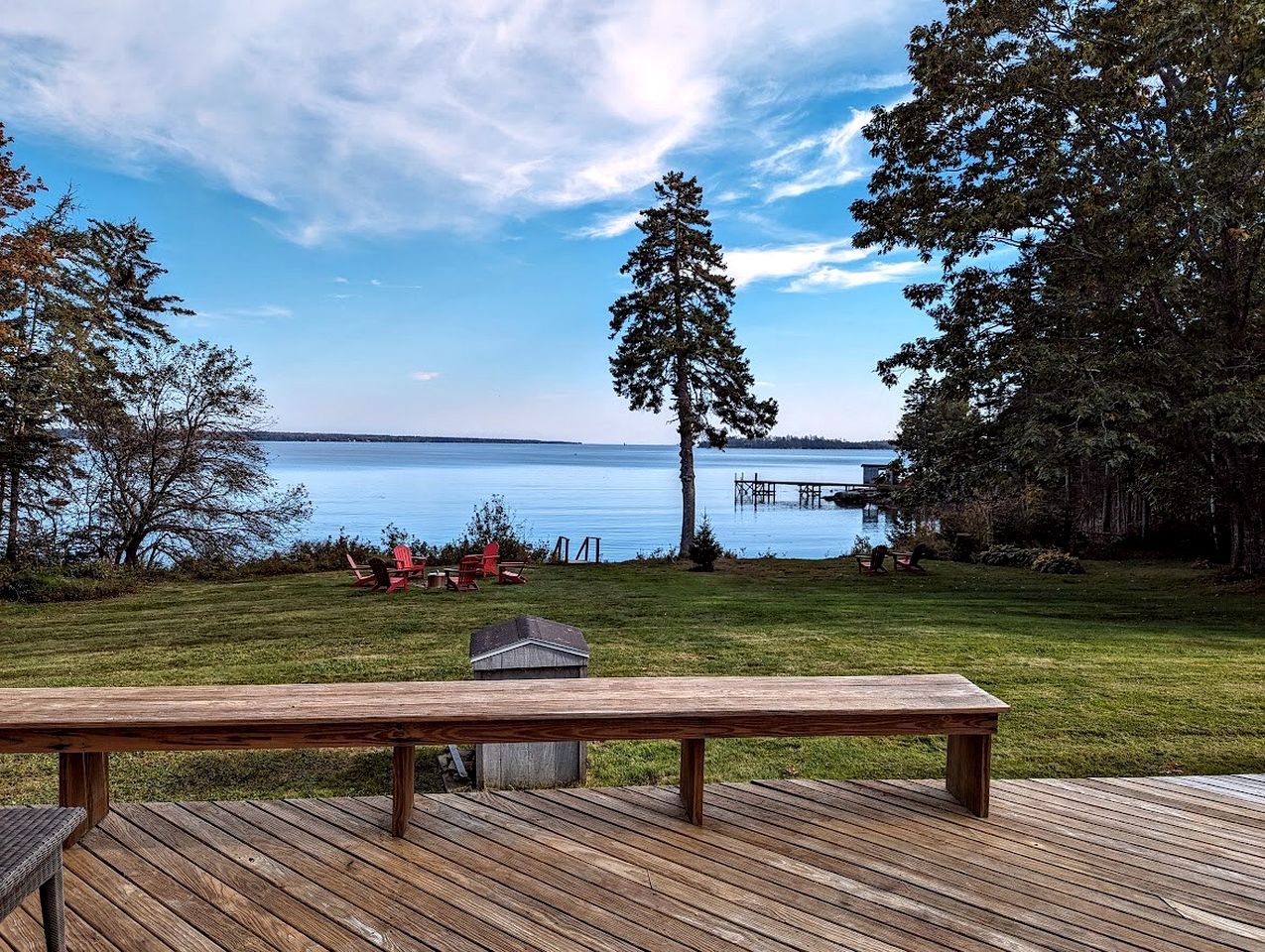 Eine Holzterrasse mit einer Bank, einem kleinen Schuppen und Blick auf einen See mit Bäumen und einem Steg.