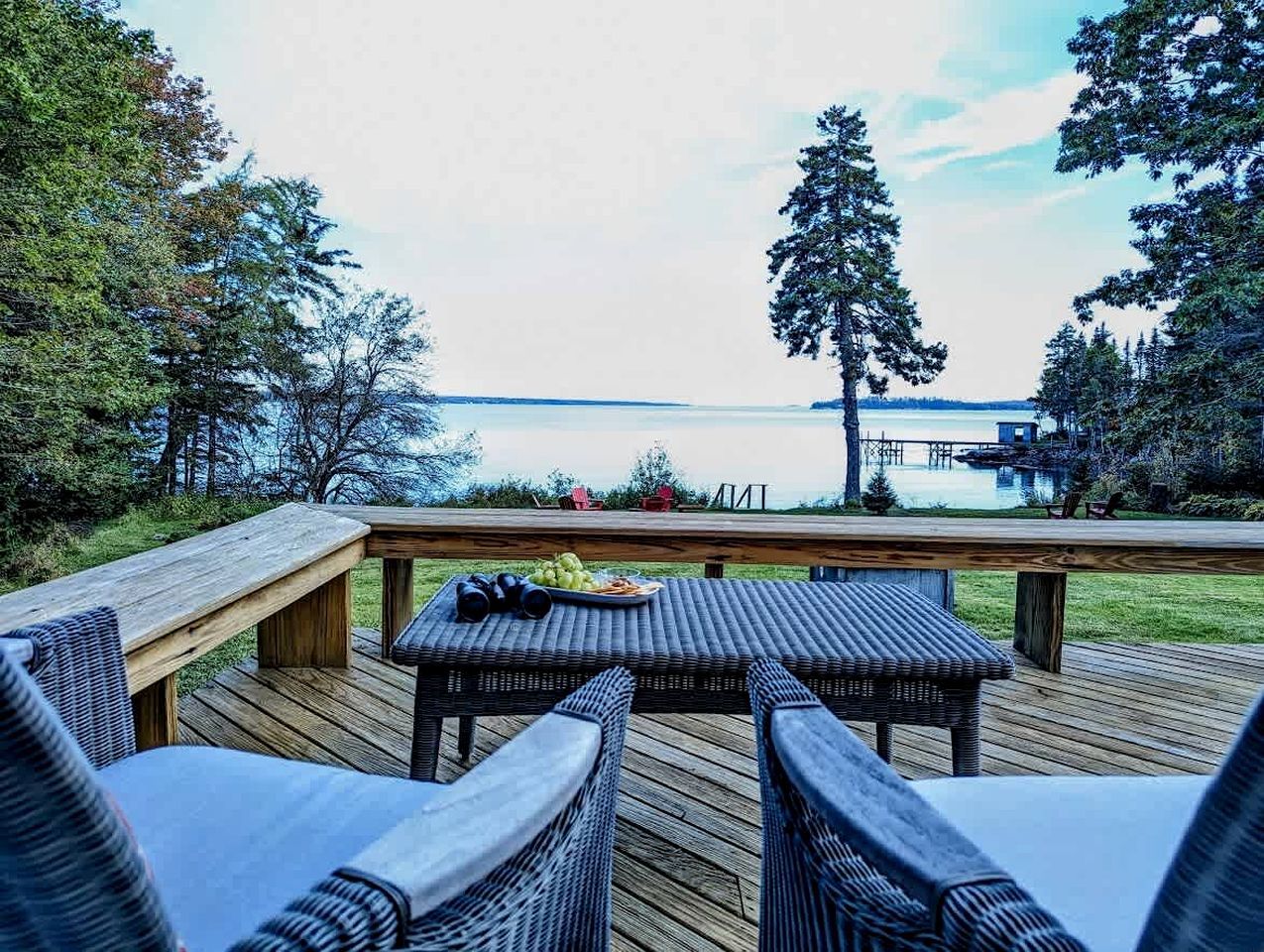 Terraza de madera con mobiliario de mimbre y una mesa, con vista a un lago que tiene un muelle y árboles.