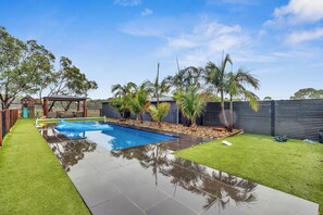 Un jardín trasero con piscina, césped artificial, palmeras y un área cercada.
