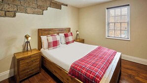 Ein Schlafzimmer mit einem hölzernen Kopfteil, einem Bett mit rotem Karomuster, zwei Nachttischlampen, einem Holznachttisch und einem Fenster mit Jalousien.