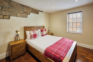 A bedroom with a wooden headboard, a bed with a red checkered blanket, two bedside lamps, a wooden nightstand, and a window with blinds. - Host & Stay | Hidden Winds Cottage (Guisborough)
