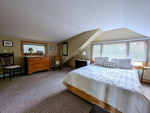 A bedroom with a sloped ceiling, a bed with a white bedspread, a wooden dresser, a chair, a nightstand with a lamp, and a window with blinds. - Oceanfront Cottage with Fire Pit & Direct Water Access Near Acadia National Park in Hancock, Maine (Hancock)