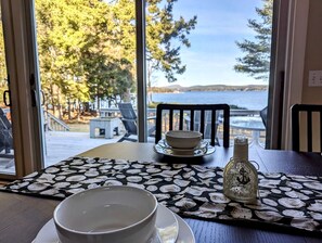 A dining table with a placemat, a bowl, a plate, a bottle, and a view of a lake through large windows. - Oceanfront Cottage with Fire Pit & Direct Water Access Near Acadia National Park in Hancock, Maine (Hancock)