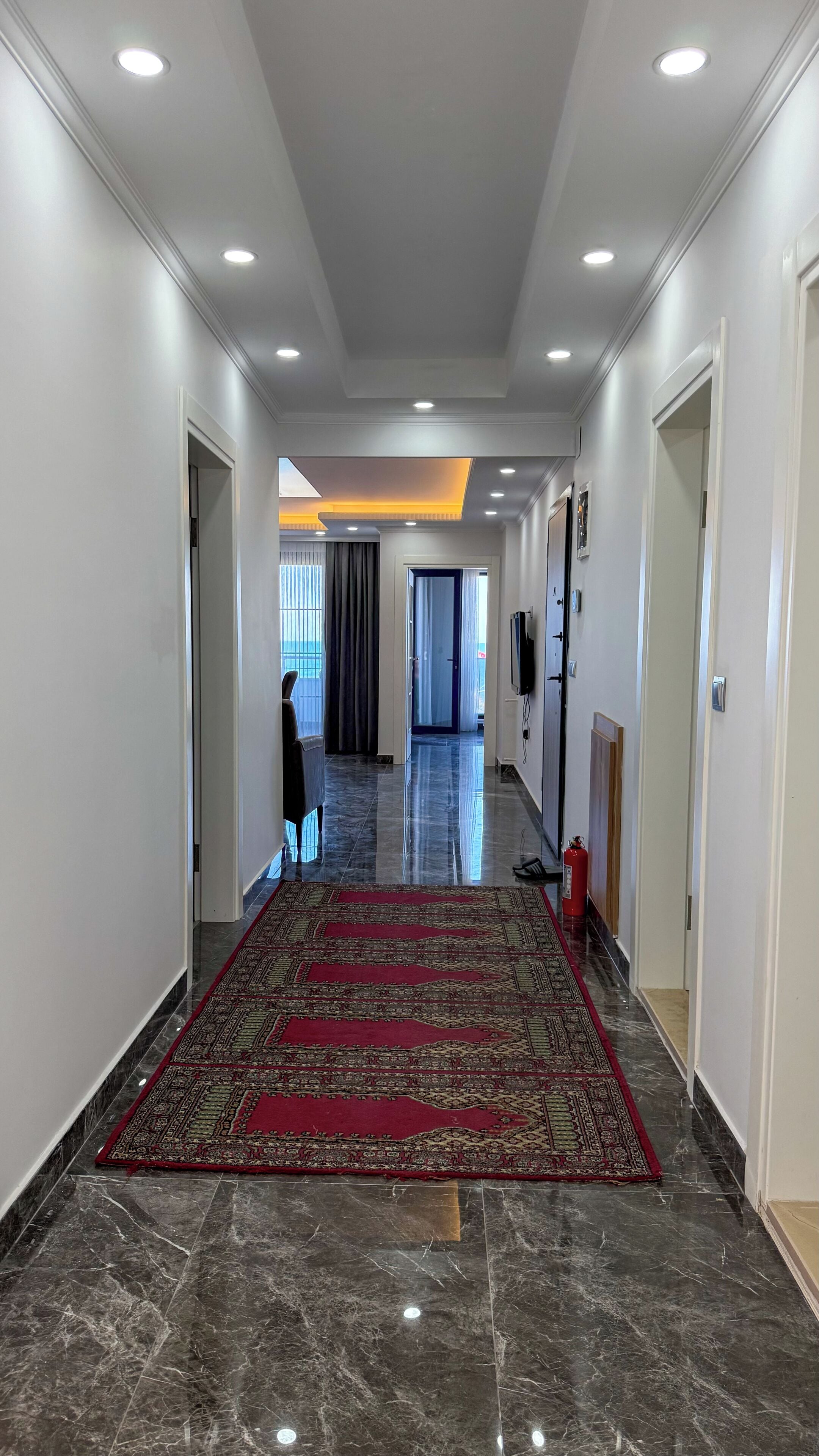 A hallway with a red patterned carpet, marble flooring, recessed ceiling lights, and a television mounted on the wall.
