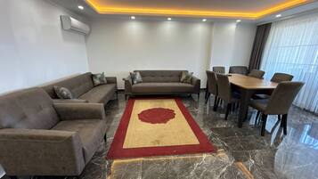 A modern living room with a sofa set, a dining table with chairs, a red and beige rug, and a large window with curtains.
