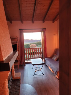A room with a wooden ceiling, a balcony with a view of a green landscape, a sofa, and a table. - A peaceful mountain chalet at an elevation of 800 meters (Sapois)