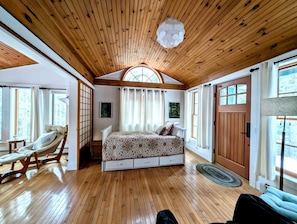 Ein Schlafzimmer mit Holzbalkendecke, einem Bett mit gemusterten Textilien, einem Sessel und einem großen Fenster.