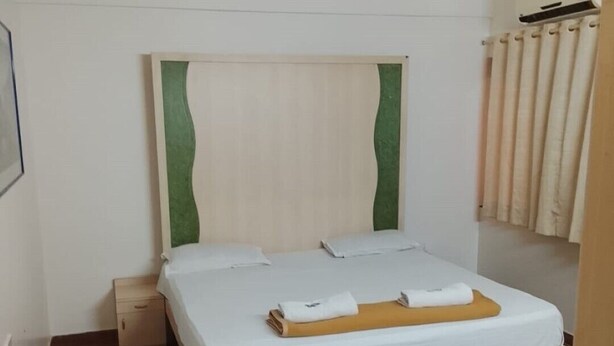 A single bed with a wooden frame and white bedding, a small bedside table, a ceiling fan, and a window with curtains. - Hotel De Rajnigandha (Nashik)