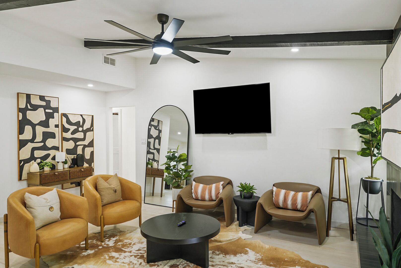 Ein modernes Wohnzimmer mit Deckenventilator, Flachbildfernseher, zwei Ledersofas mit gemusterten Kissen, einem runden Couchtisch und einem Kuhfellteppich.