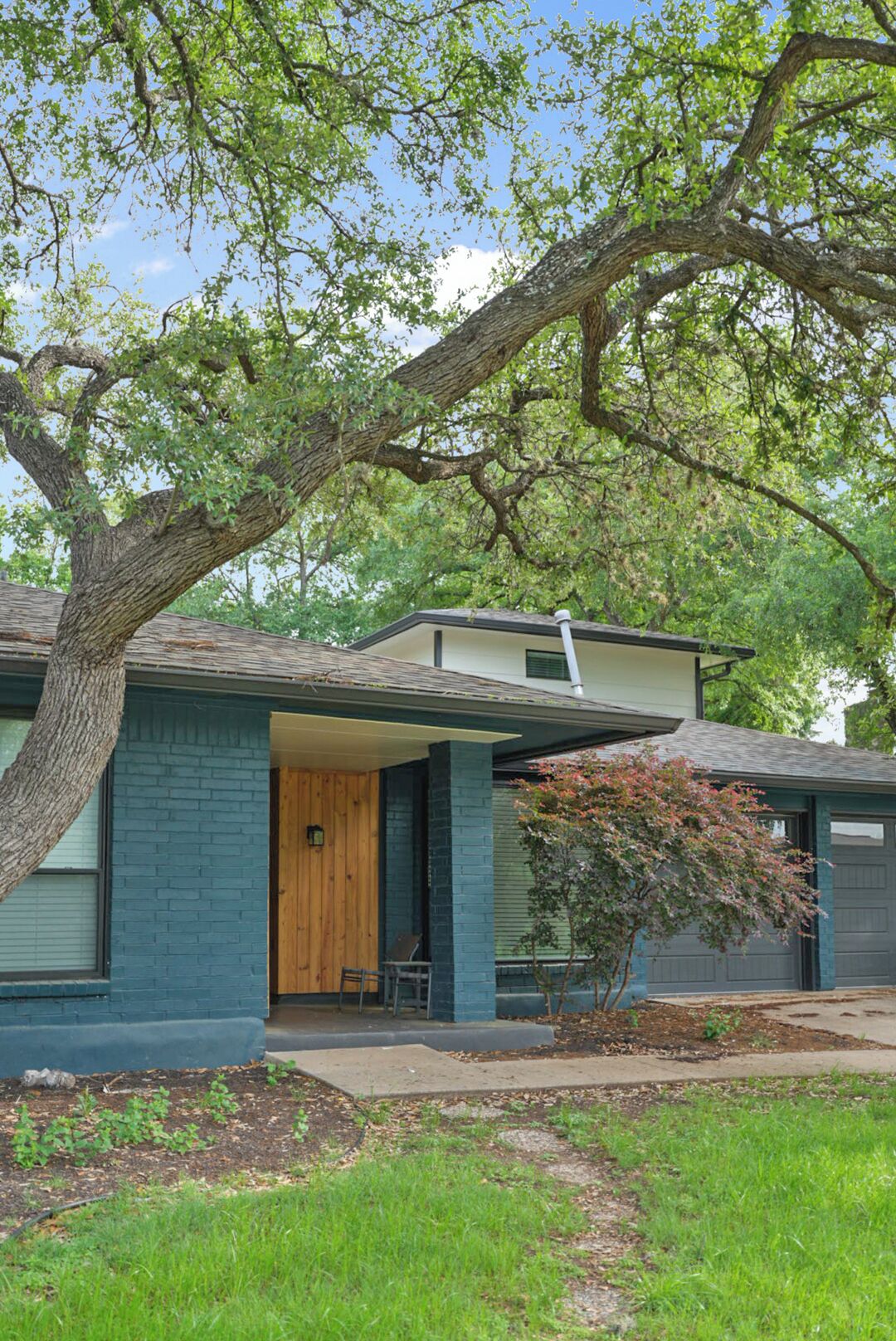 A residential house with a front porch, a garage, and a large tree in the yard.