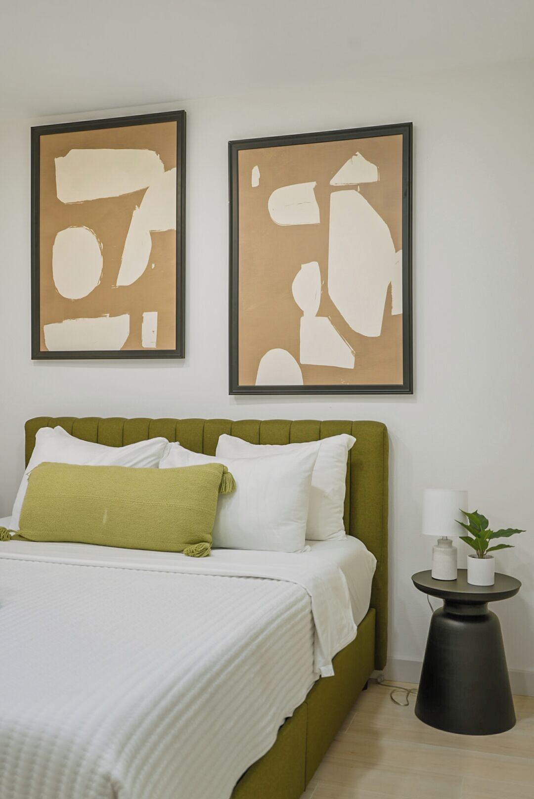 Un dormitorio con un cabecero tapizado en verde, ropa de cama blanca, dos cuadros abstractos en la pared y una mesita de noche con una lámpara y una planta.