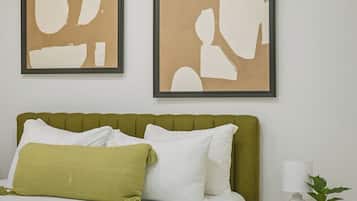 Un dormitorio con un cabecero tapizado en verde, ropa de cama blanca, dos cuadros abstractos en la pared y una mesita de noche con una lámpara y una planta.