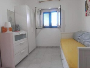 Ein kleines Zimmer mit Bett, Schreibtisch, Fenster mit Jalousien und einem Schrank.