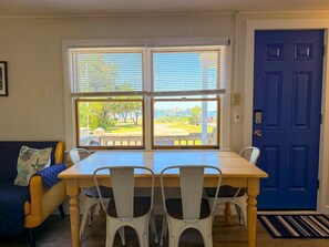 A dining area with a table and chairs, a blue door, and a view of a park through large windows. - Ocracoke Local 5: Close to Harbor | Private Boat Slip | Queen Bed (Ocracoke)