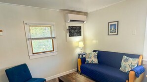 Een woonkamer met een blauwe bank, een blauwe fauteuil, een houten salontafel, een wandairconditioner, een raam met jaloezieën, een kleine tafel met een lamp en een ingelijst schilderij aan de muur.