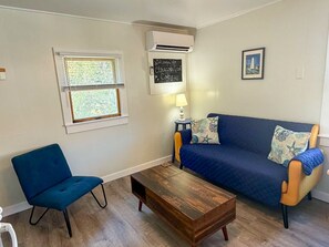 A living room with a blue sofa, a blue chair, a wooden coffee table, a wall-mounted air conditioner, a window with blinds, a small table with a lamp, and a framed picture on the wall. - Ocracoke Local 5: Close to Harbor | Private Boat Slip | Queen Bed (Ocracoke)