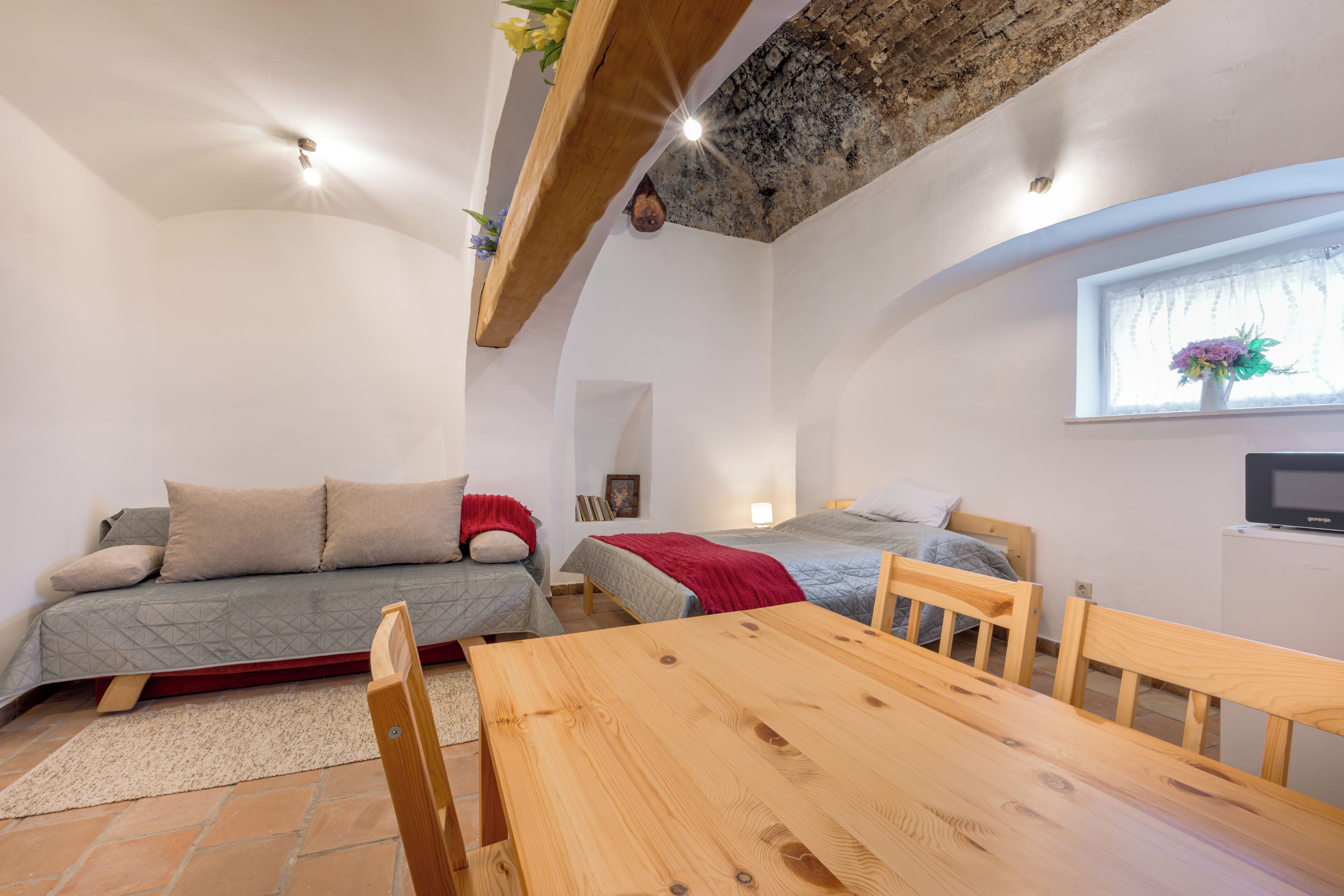 A compact room with a wooden table, two beds, a sofa, and a window with a flower arrangement.
