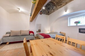 A compact room with a wooden table, two beds, a sofa, and a window with a flower arrangement.