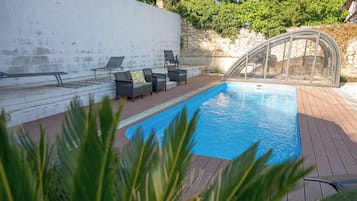 A swimming pool with a wooden deck, surrounded by lounge chairs and a glass enclosure.