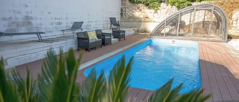 A swimming pool with a wooden deck, surrounded by lounge chairs and a glass enclosure.