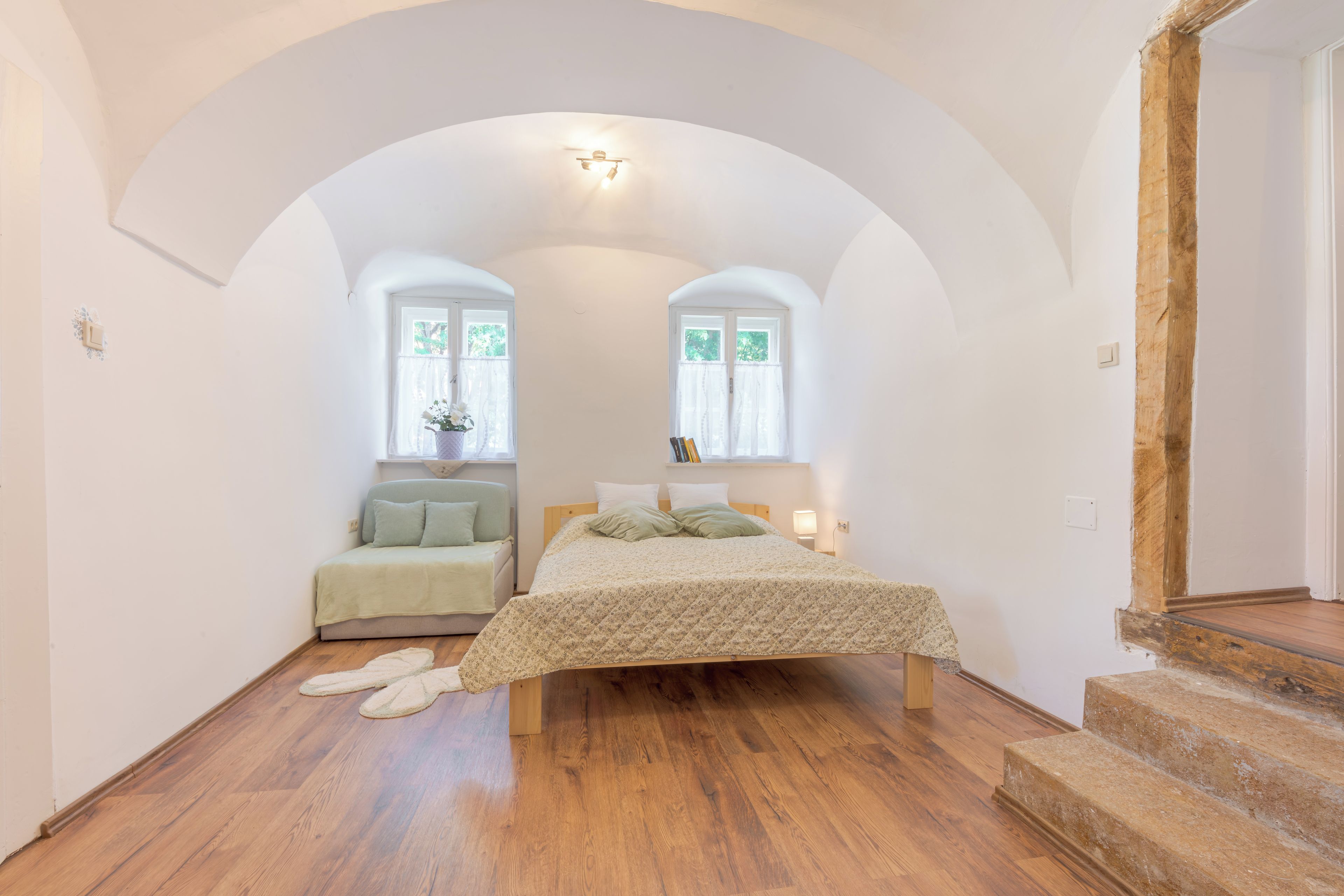 A bedroom with a wooden floor, a bed with a patterned bedspread, a nightstand, and two windows with curtains.