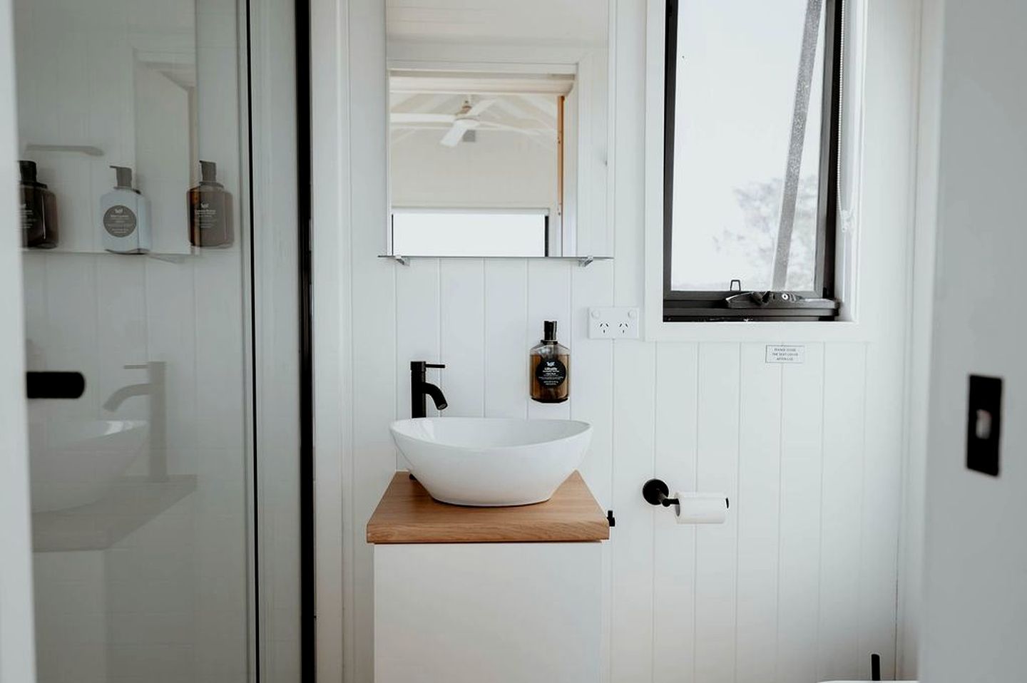 Une salle de bain moderne avec un lavabo sur colonne blanc, un robinet noir et une étagère en bois.
