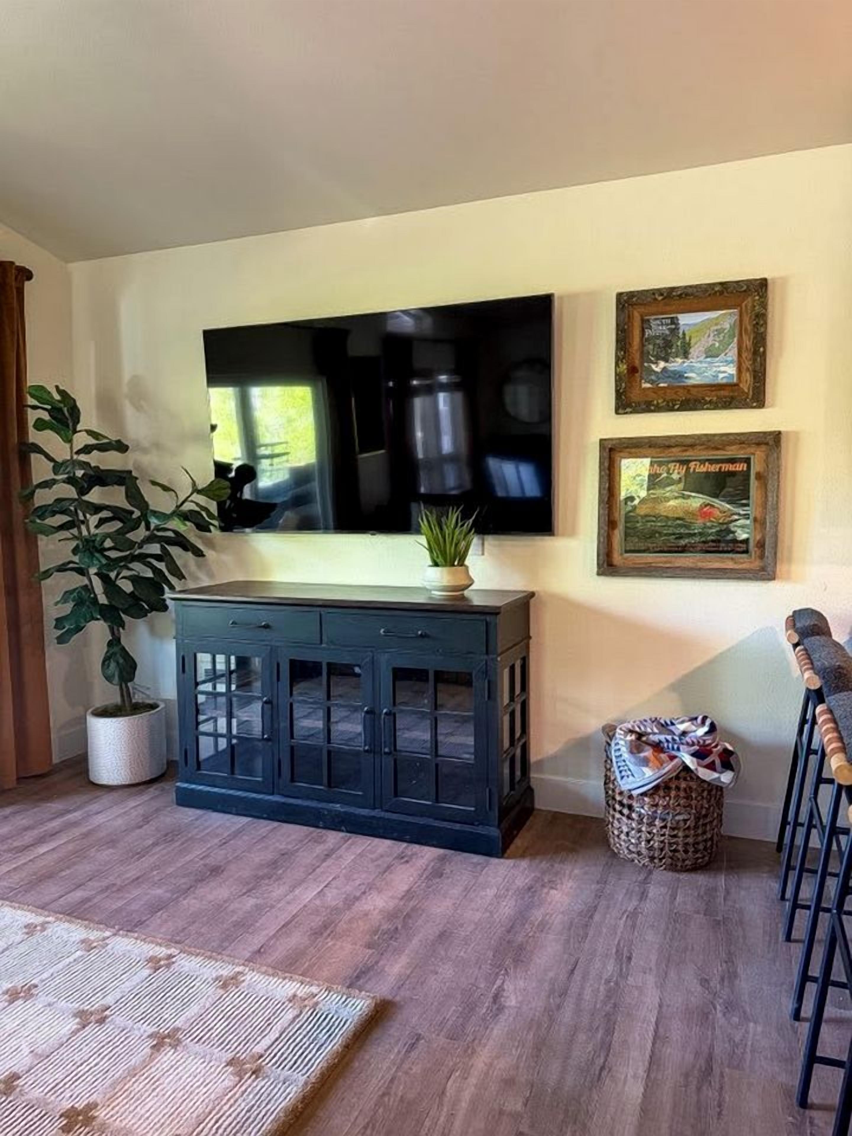 Una sala de estar con un aparador azul oscuro, un televisor de pantalla plana montado en la pared, una planta en maceta, un cuadro enmarco y una cesta tejida.