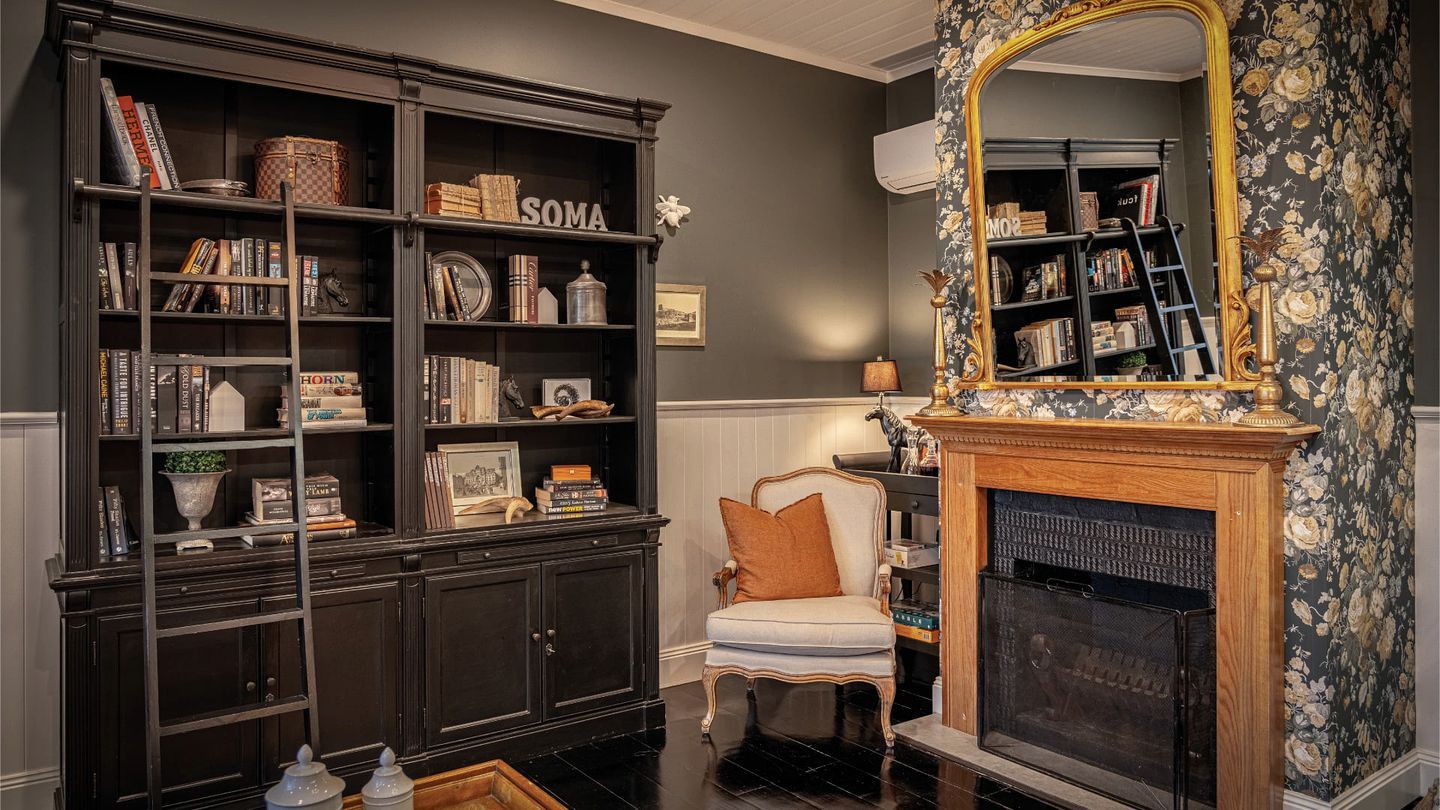 A room with a dark wood bookcase, a white armchair, a fireplace, and a large mirror with a floral frame.