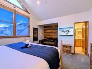Ein Hotelzimmer mit einem großen Bett, Blick auf eine Berglandschaft durch das Fenster, einer Holzwand, einem kleinen Stuhl und einer Tür zu einem anderen Raum.