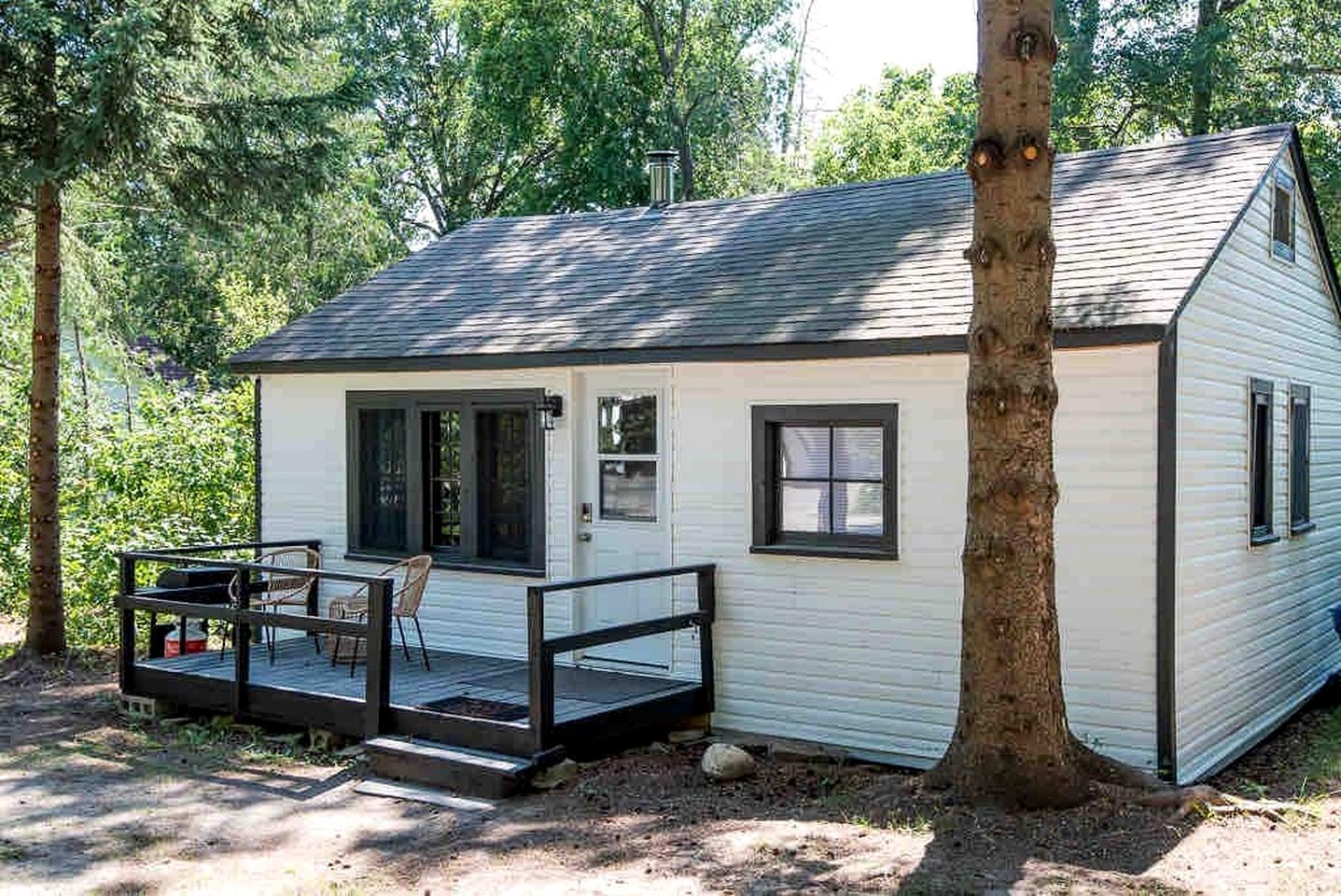 Een klein, gelijkvloers huisje met een veranda en terras, omgeven door bomen.