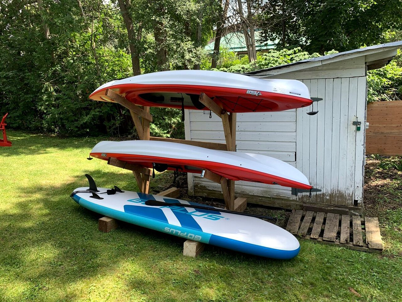 Trois planches de surf sont empilées sur une structure en bois, dans un espace herbeux, avec un abri en arrière-plan.