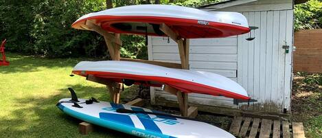 Trois planches de surf sont empilées sur une structure en bois, dans un espace herbeux, avec un abri en arrière-plan.