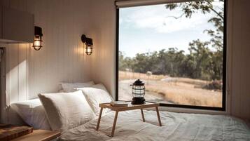 A bedroom with a bed, pillows, a bedside table, a lantern, and a view of a natural landscape through a large window.