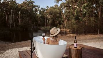 A person relaxing in a large white bathtub outdoors, surrounded by trees and a wooden deck.